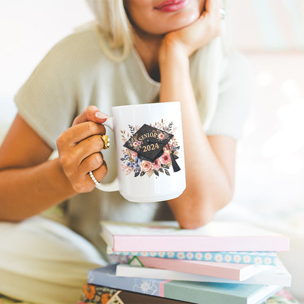 Woman holding a 15oz coffee mug that features a floral senior 2024 cap and tassel. This gift is the perfect graduation gift for her. All SKUs.