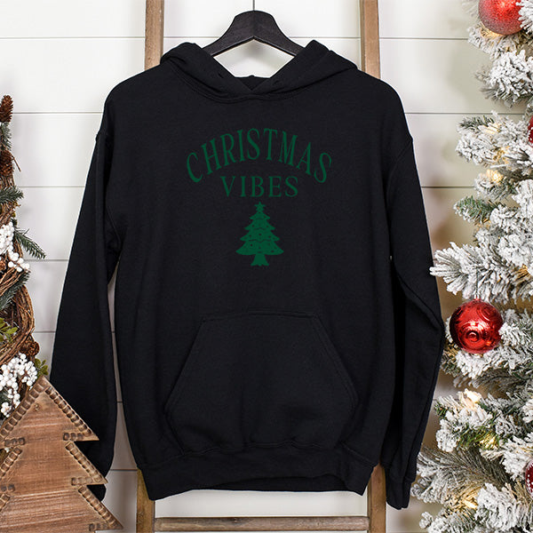 Black Christmas hoodie hanging with Christmas tree in the background. This unisex hoodie has green lettering showing Christmas Vibes and a Christmas tree. all SKUs