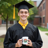 Male graduate wearing a black cap and gown, standing outdoors and holding a Class of 2025 mug with both hands, smiling as he celebrates his achievement. Colorful graduation mug with male graduate. 