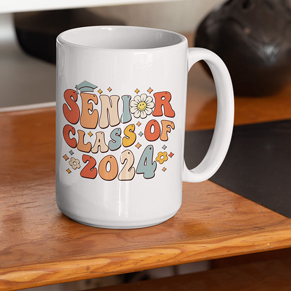 Senior class of 2024 graduation coffee mug sitting on a wooden desk, next to a computer. A great grad gift idea for her. All SKUs. 