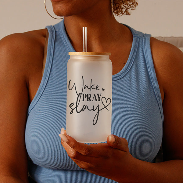 Woman holding Frosted 16oz Glass Tumbler with black Wake, Pray, Slay design. This motivational glassware comes with a bamboo lid and a straw.