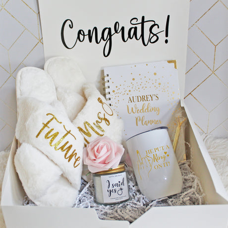 Gold themed engagement gift bundle for future mrs. Includes a pair of fluffy slippers, a custom wine tumbler, a personalized wedding planner, jewel pen and scented candle.