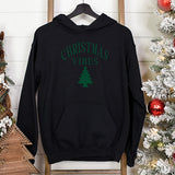 Black Christmas hoodie hanging with Christmas tree in the background.  This unisex hoodie has green lettering showing Christmas Vibes and a Christmas tree.  all SKUs